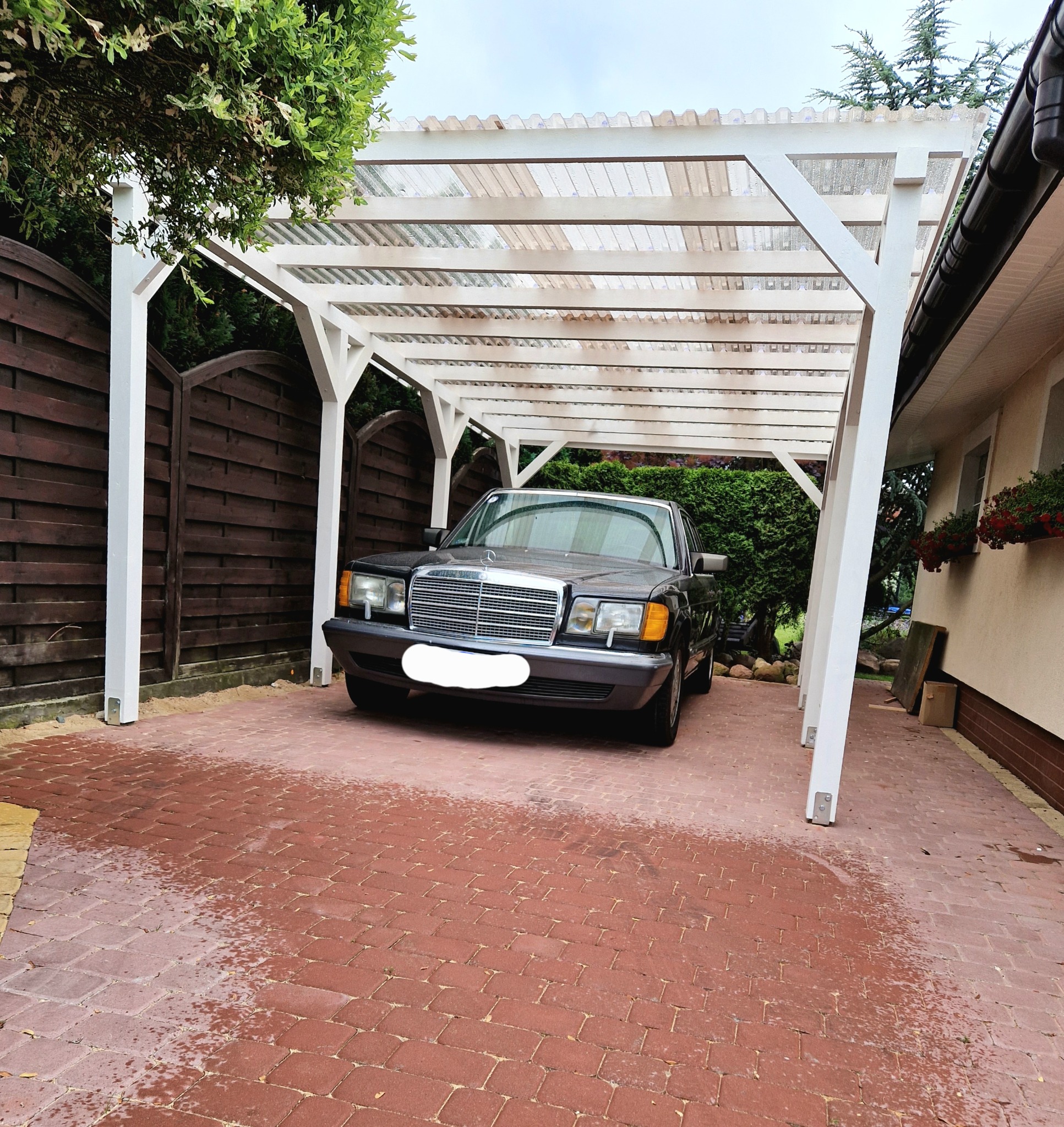 Czarny samochód osobowy zaparkowany pod białą, drewnianą konstrukcją z przezroczystym zadaszeniem, obok domu z czerwonymi kwiatami w oknach, na podjeździe z czerwonej kostki brukowej.