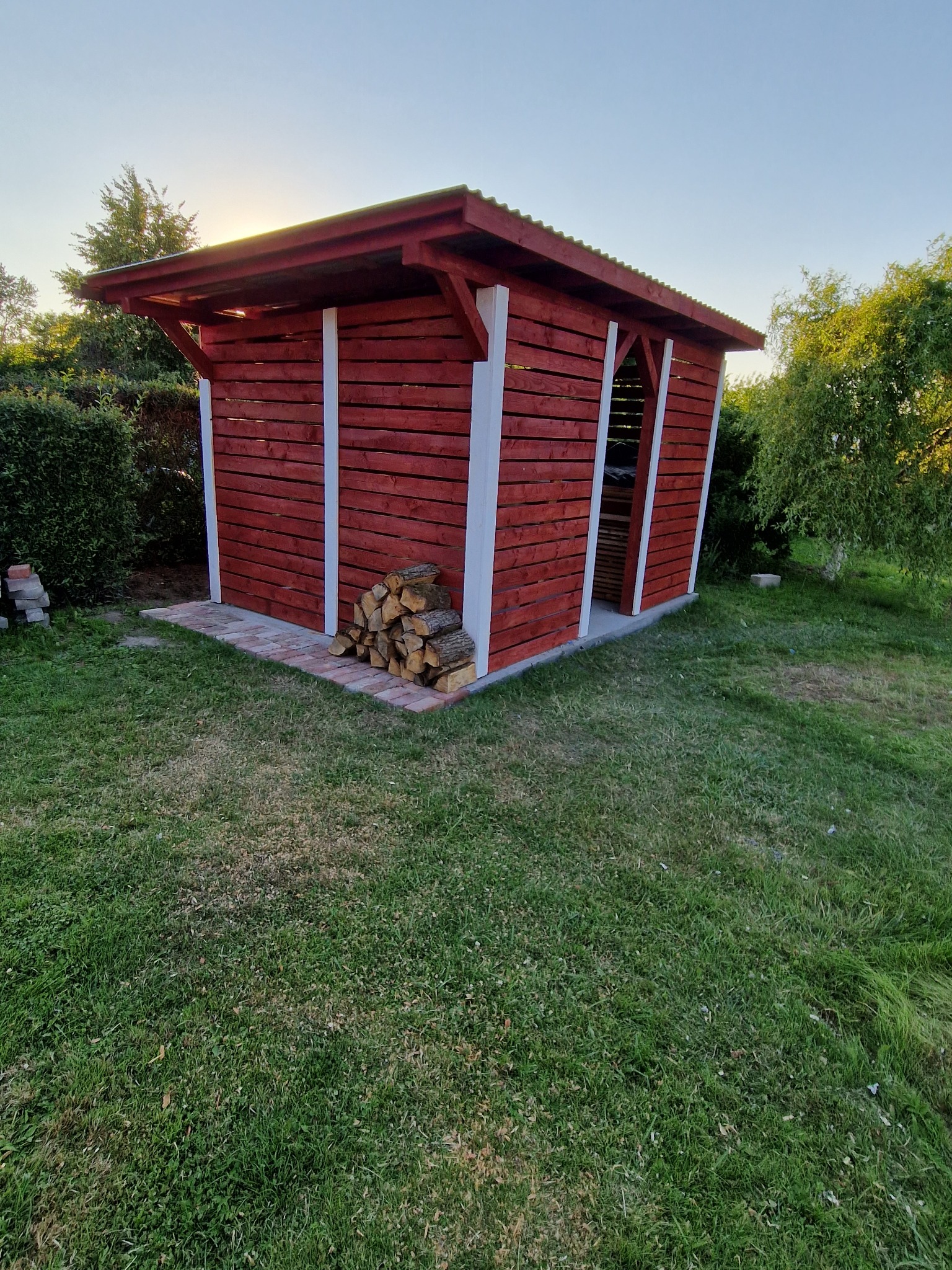 Czerwona drewniana altana na drewno z białymi detalami i stosem drewna, otoczona zielonym trawnikiem i krzewami.