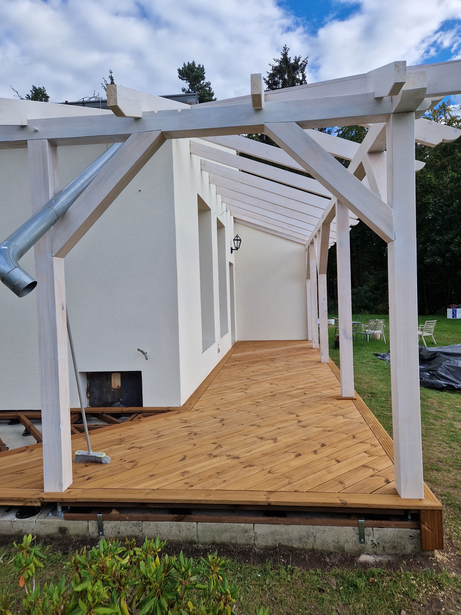 Biała drewniana konstrukcja zadaszenia tarasu w trakcie budowy, z widoczną nową drewnianą podłogą, fragmentem elewacji budynku z rynną i szczotką opartą o filar.