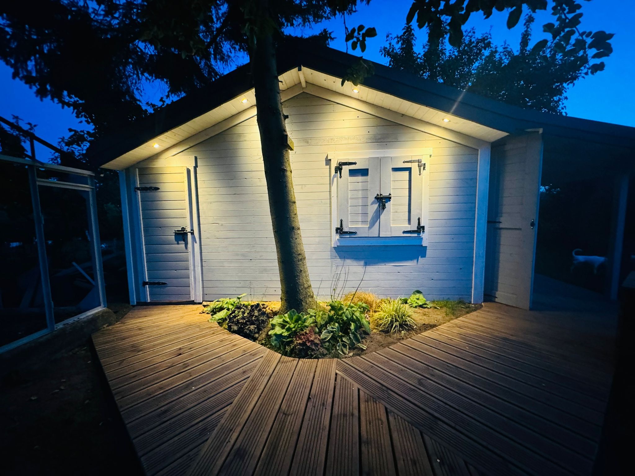 Oświetlona altana z białymi ścianami i drewnianym tarasem otaczającym drzewo, widoczna wieczorem z podświetleniem punktowym.