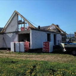 Budowa domu z betonu komórkowego w toku, widoczne palety z materiałem Termobet, konstrukcja dachu wzniesiona, w tle krajobraz górski i błękitne niebo.