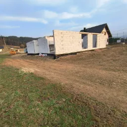 Wznoszenie ścian parterowego domu z białych bloczków, widoczne otwory okienne i drzwiowe. W tle las i częściowo widoczny budynek z ciemnym dachem.