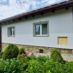 Biała elewacja domu jednorodzinnego z oknami w jasnych ramach, widoczny fragment kamiennej podmurówki i klapa gazowa, ogród z bujną roślinnością na pierwszym planie.