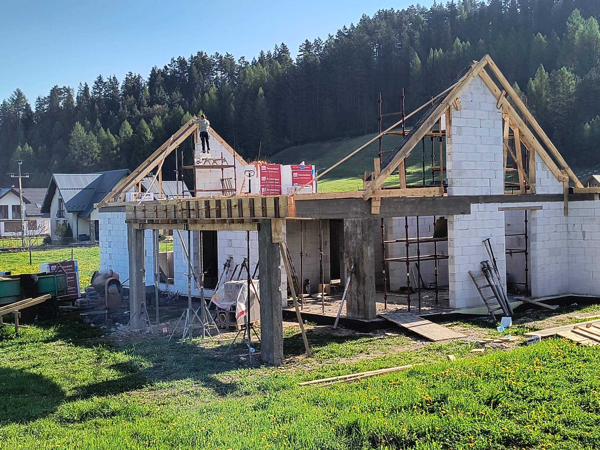 Wznoszenie ścian z bloczków na budowie domu jednorodzinnego w słoneczny dzień. Widoczne rusztowania, drewniane elementy konstrukcyjne i pracownik na dachu.