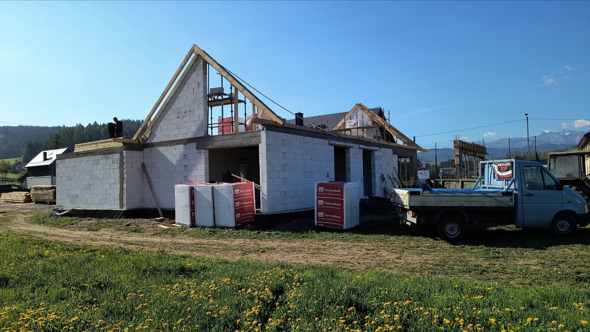 Budowa domu z betonu komórkowego w toku, widoczne palety z materiałem Termobet, konstrukcja dachu wzniesiona, w tle krajobraz górski i błękitne niebo.