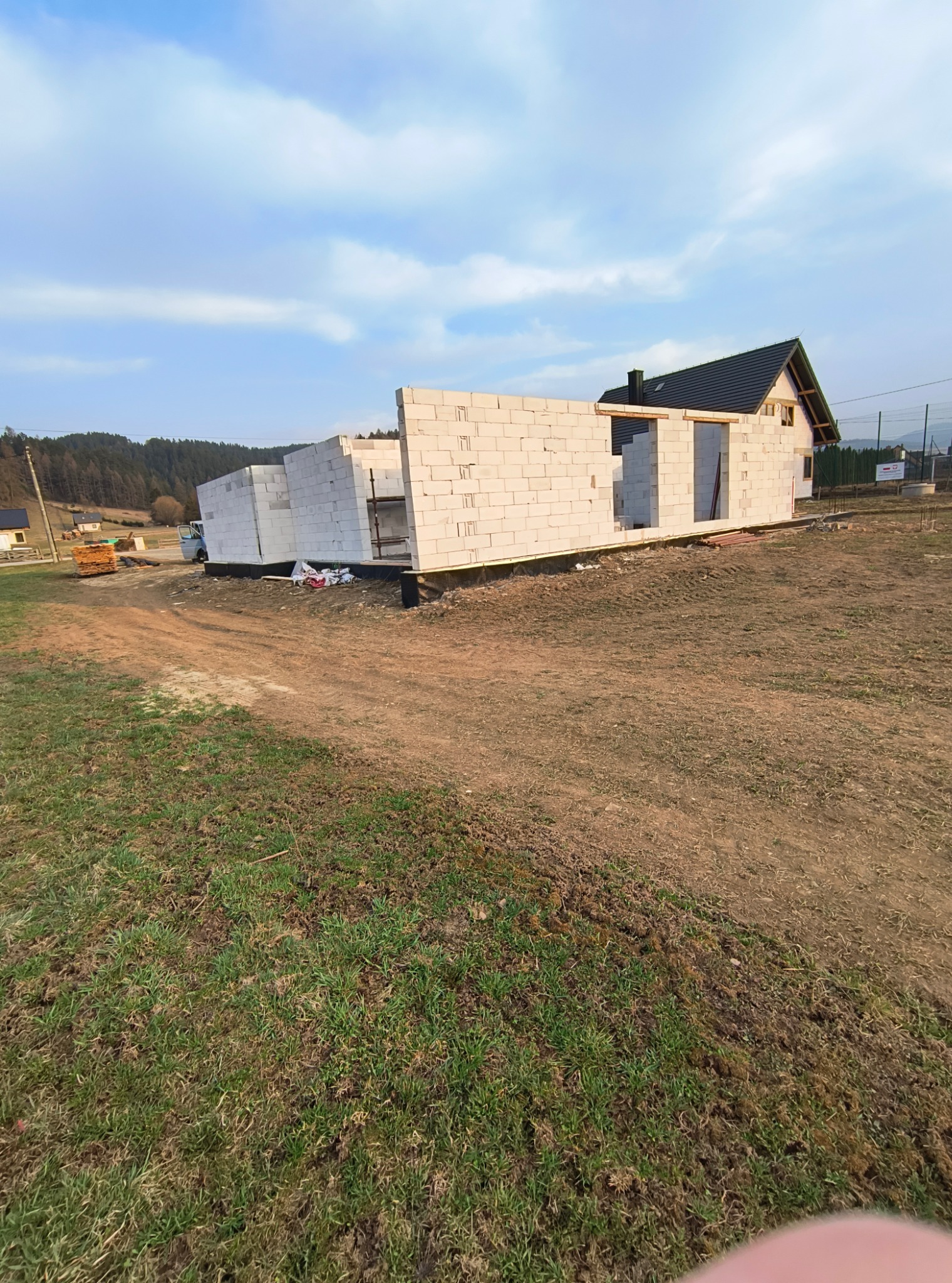 Wznoszenie ścian parterowego domu z białych bloczków, widoczne otwory okienne i drzwiowe. W tle las i częściowo widoczny budynek z ciemnym dachem.