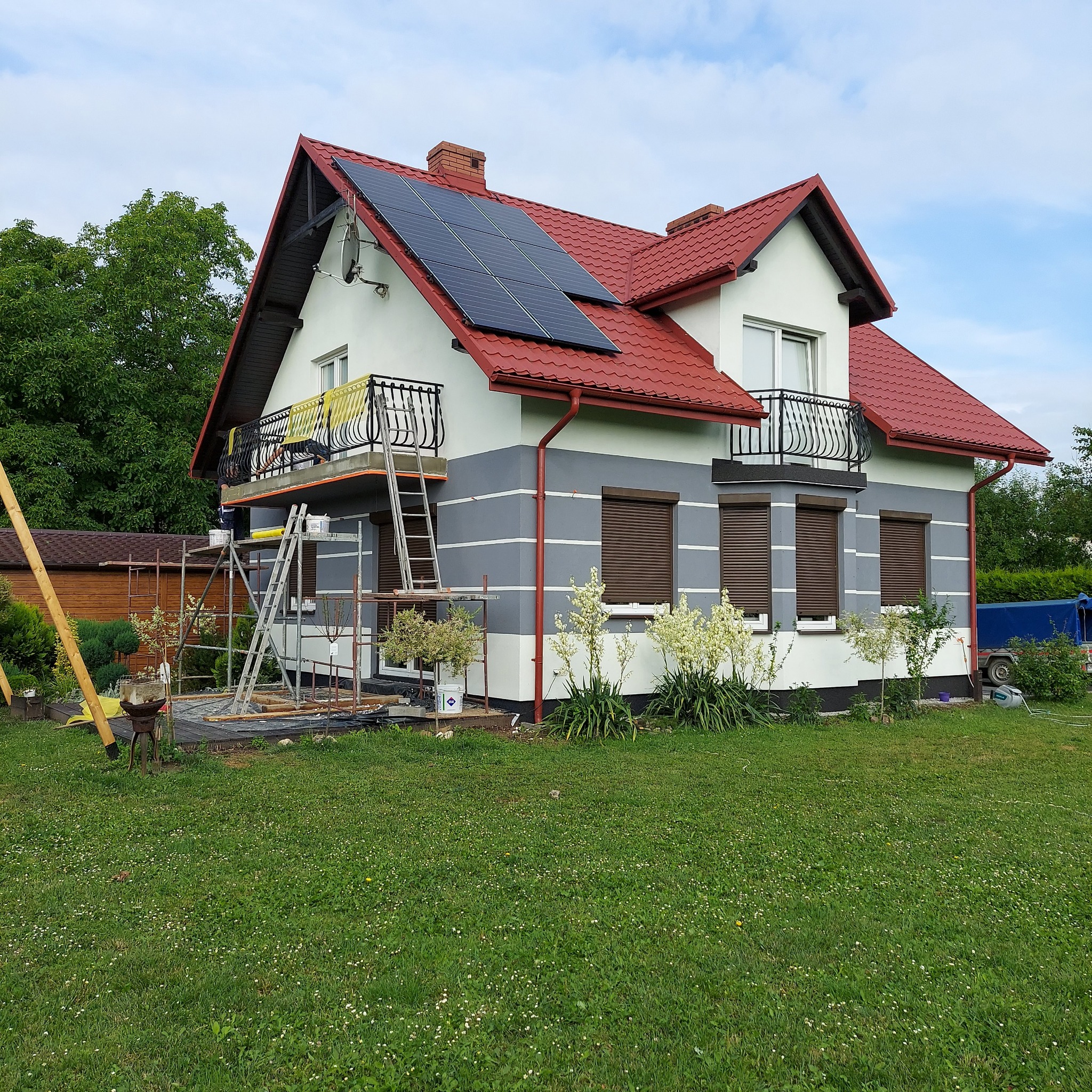 Dwukondygnacyjny dom z czerwonym dachem i panelami słonecznymi na etapie prac elewacyjnych, widoczne rusztowania i drabina przy balkonie.