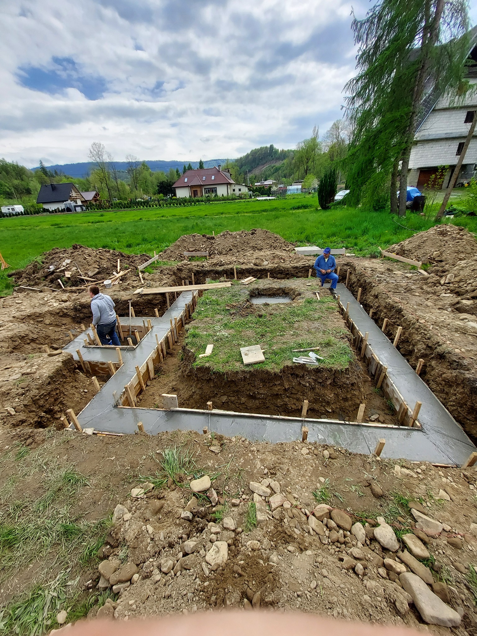 Budowa fundamentów domu: zalewanie betonem szalunków, widoczni dwaj pracownicy w odzieży roboczej, krajobraz wiejski w tle.