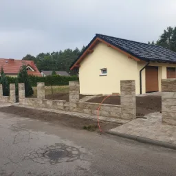 Wznoszenie ogrodzenia z elementów betonowych imitujących kamień, z widoczną podbudową i słupkami, na tle żółtego budynku i kostki brukowej.