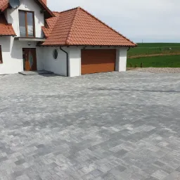 Nowoczesny dom jednorodzinny z szarą kostką brukową ułożoną we wzór jodełki na podjeździe, z widokiem na zielone pola w tle.