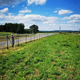 Mężczyzna w czapce i rękawicach naciąga siatkę na słupki nowego ogrodzenia z siatki, widok na łąkę i błękitne niebo z chmurami.
