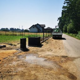 Xenimar - Budowa ogrodzenia: betonowe słupki z wystającymi prętami zbrojeniowymi, podmurówka w trakcie wylewania, w tle zielony traktor na pace samochodu dostawczego, widok na dom i pole.