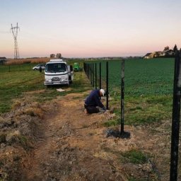 Xenimar - Pracownik w trakcie montażu słupków ogrodzeniowych na polu, widoczny biały samochód dostawczy i ciągnik rolniczy w tle, linia energetyczna w oddali.