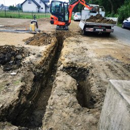 Xenimar - Głęboki wykop w ziemi, z pomarańczową mini koparką Kubota i wywrotką wypełnioną ziemią w tle, widoczne linie wyznaczające obszar robót, w oddali budynek mieszkalny.