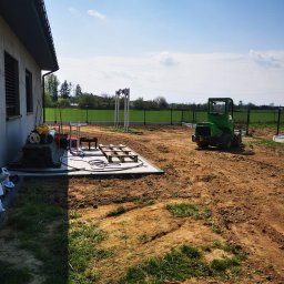 Xenimar - Przygotowany teren pod budowę domu z widocznym ogrodzeniem panelowym, małym traktorem i materiałami budowlanymi przy ścianie budynku.