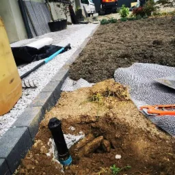 Instalacja zraszacza w ziemi przy obrzeżu trawnika, widoczna żółta beczka, grabie, łopata i nożyce ogrodowe oraz trawa z rolki w tle.