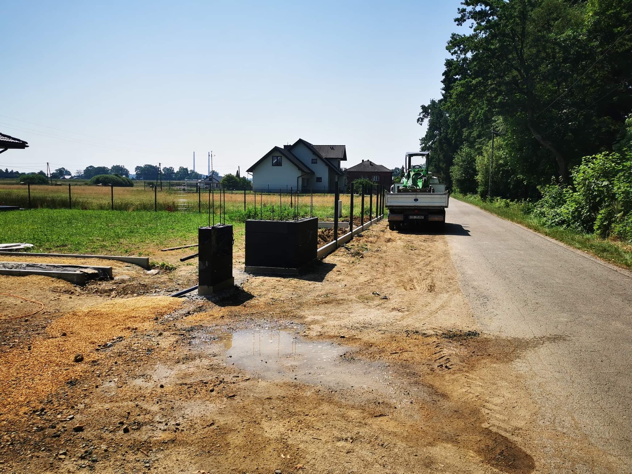 Budowa ogrodzenia: betonowe słupki z wystającymi prętami zbrojeniowymi, podmurówka w trakcie wylewania, w tle zielony traktor na pace samochodu dostawczego, widok na dom i pole.