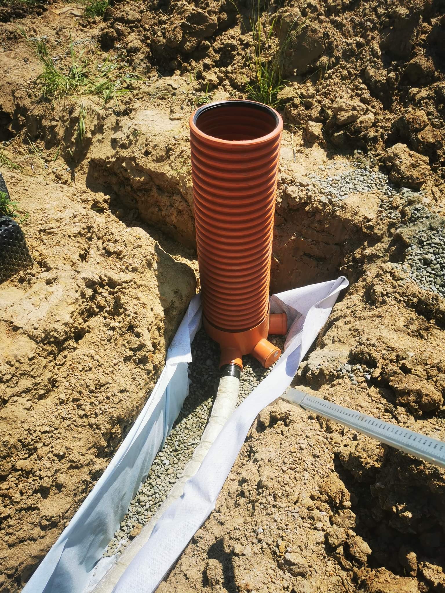 Pomarańczowa, karbowana rura pionowa z przyłączami, umieszczona w wykopie z geowłókniną i drenażem żwirowym, częściowo zasypana ziemią.