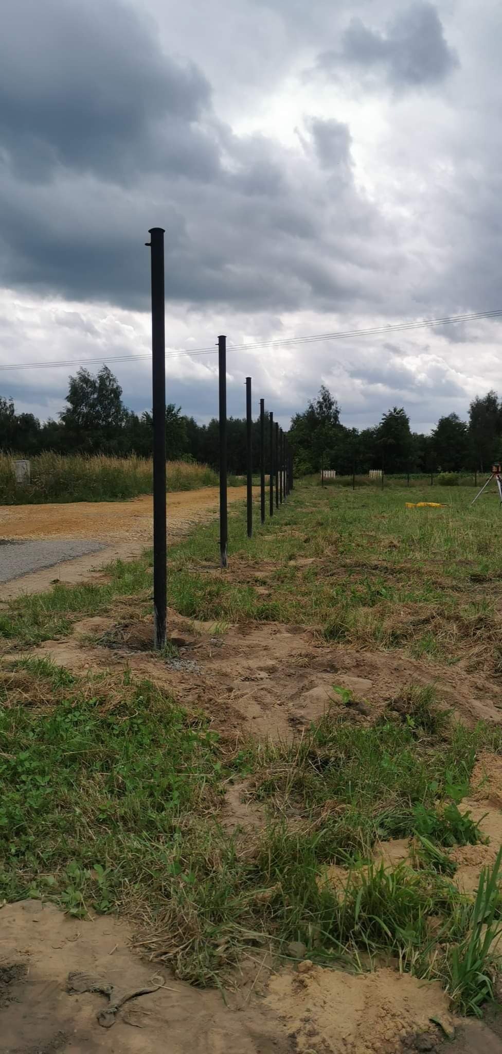 Rząd czarnych, metalowych słupków ogrodzeniowych, częściowo osadzonych w ziemi na zewnątrz, z widocznym krajobrazem polnym i pochmurnym niebem w tle.