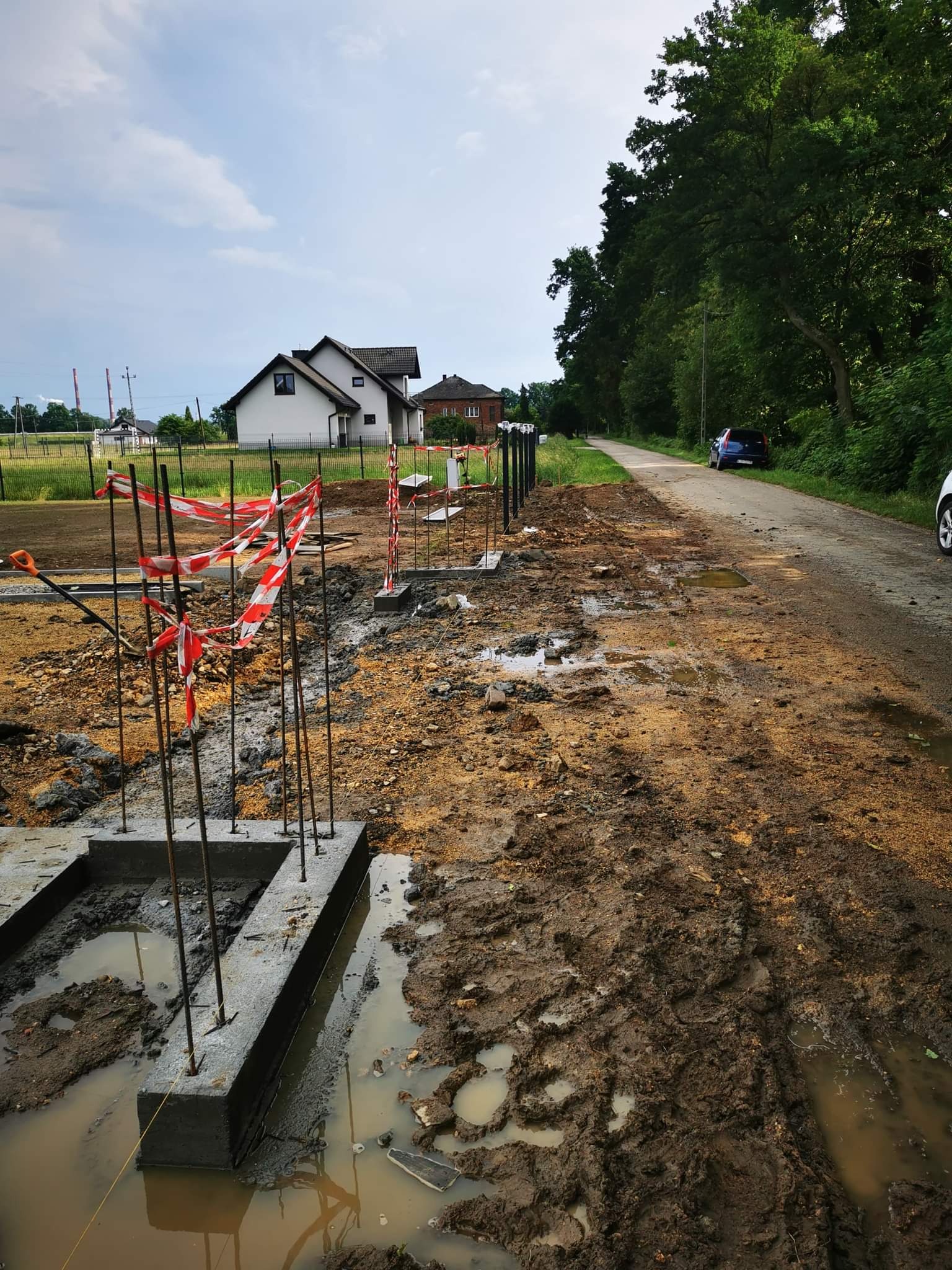 Włosienica: Wykop pod fundamenty zbrojony stalowymi prętami, zabezpieczony taśmą ostrzegawczą, w tle dom jednorodzinny i droga gruntowa.