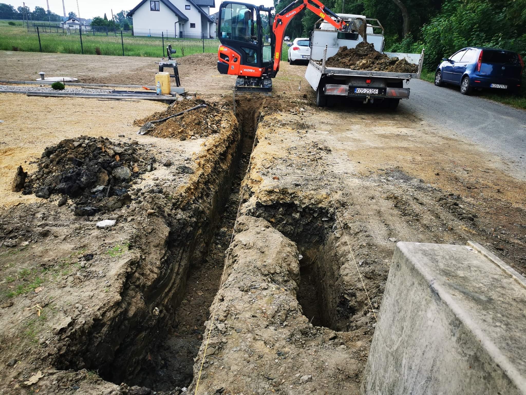 Głęboki wykop w ziemi, z pomarańczową mini koparką Kubota i wywrotką wypełnioną ziemią w tle, widoczne linie wyznaczające obszar robót, w oddali budynek mieszkalny.