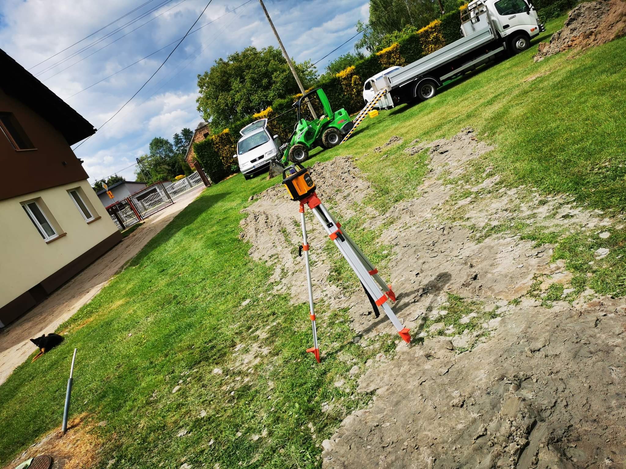 Niwelator laserowy na trójnogu ustawiony na przygotowanym terenie, w tle minikoparka, samochód dostawczy i budynek mieszkalny.