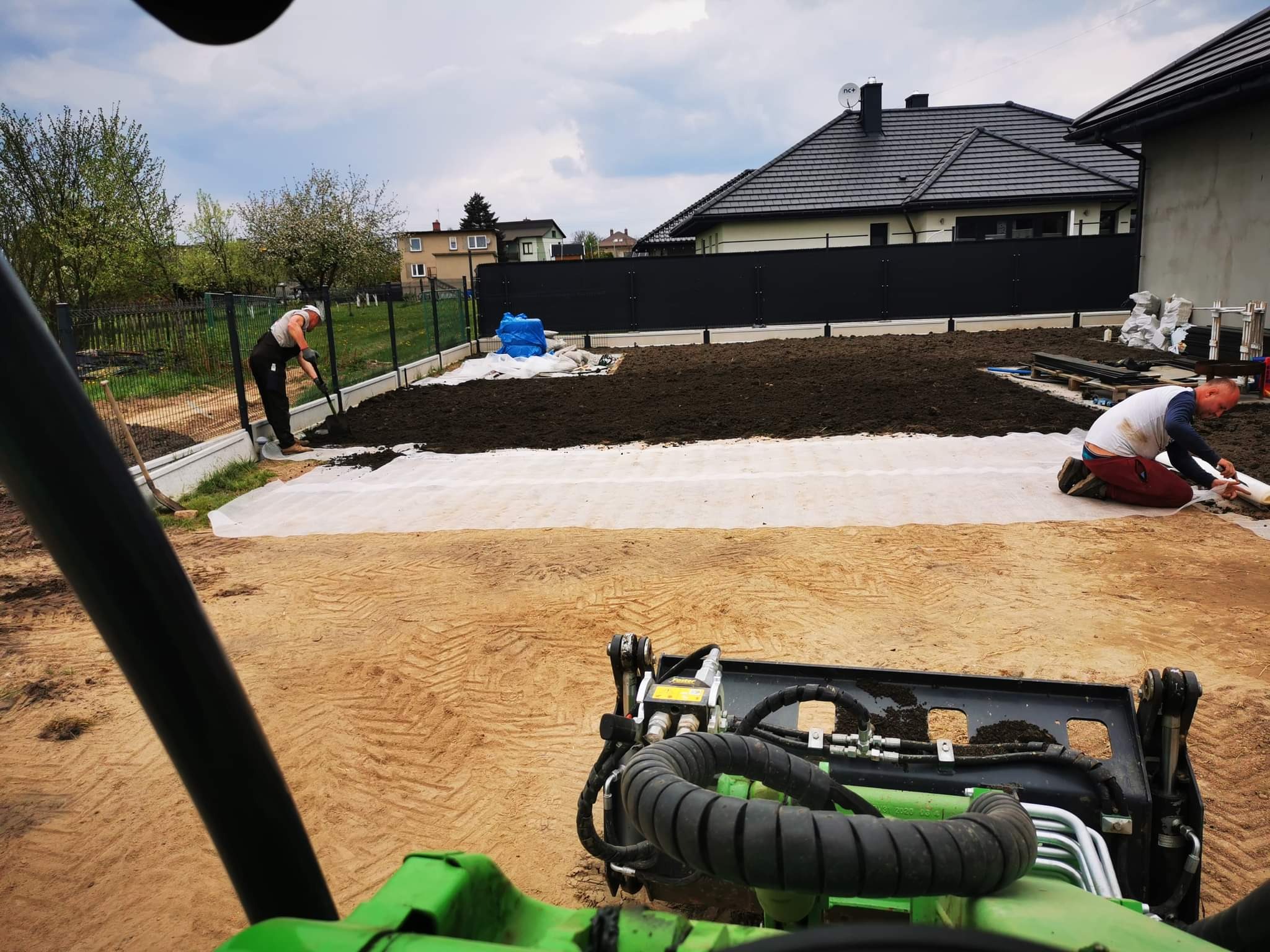 Przygotowanie terenu pod trawnik: wyrównywanie ziemi na geowłókninie, widok z kabiny miniładowarki, w tle ogrodzenie panelowe i dom