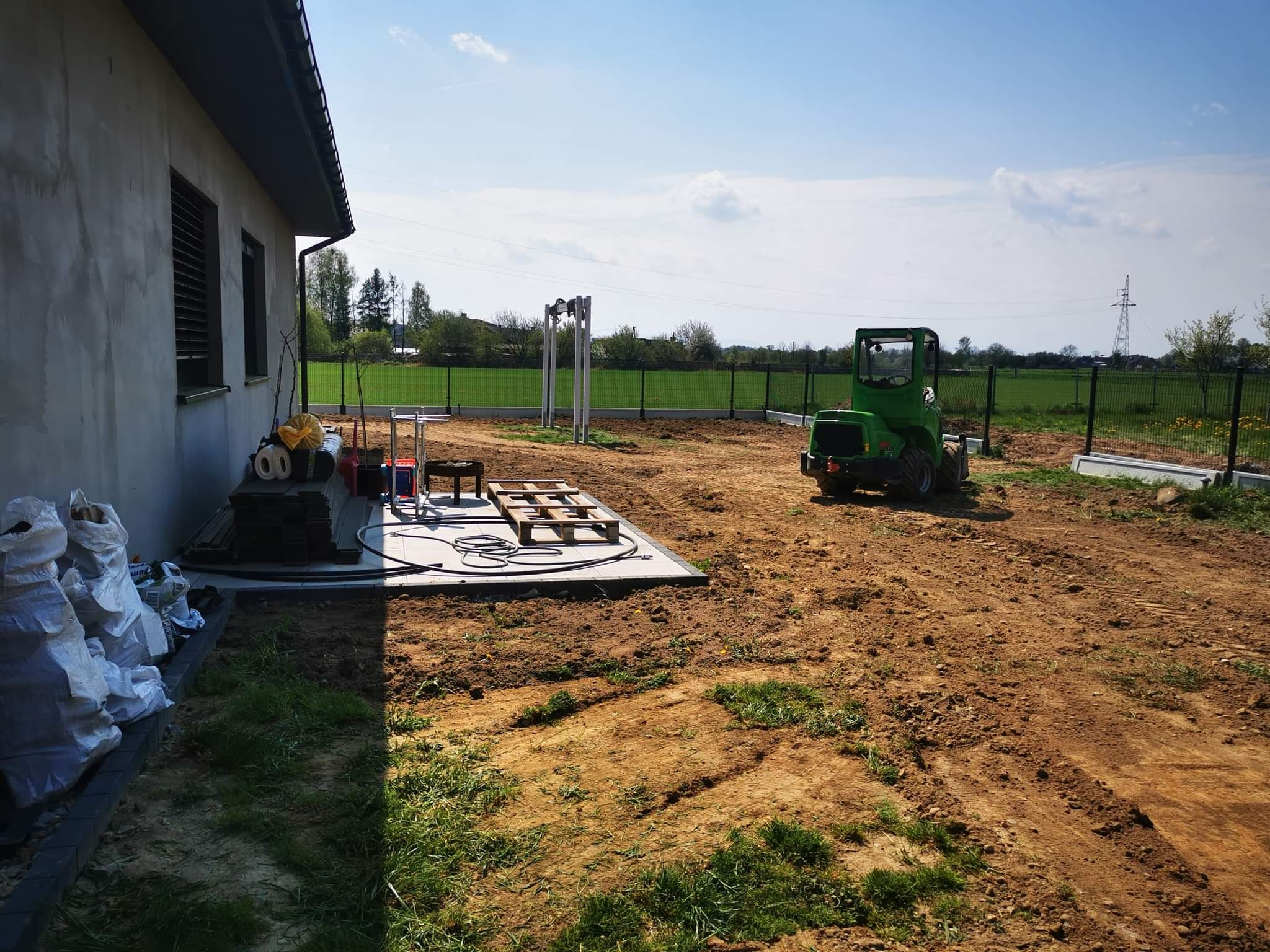 Przygotowany teren pod budowę domu z widocznym ogrodzeniem panelowym, małym traktorem i materiałami budowlanymi przy ścianie budynku.
