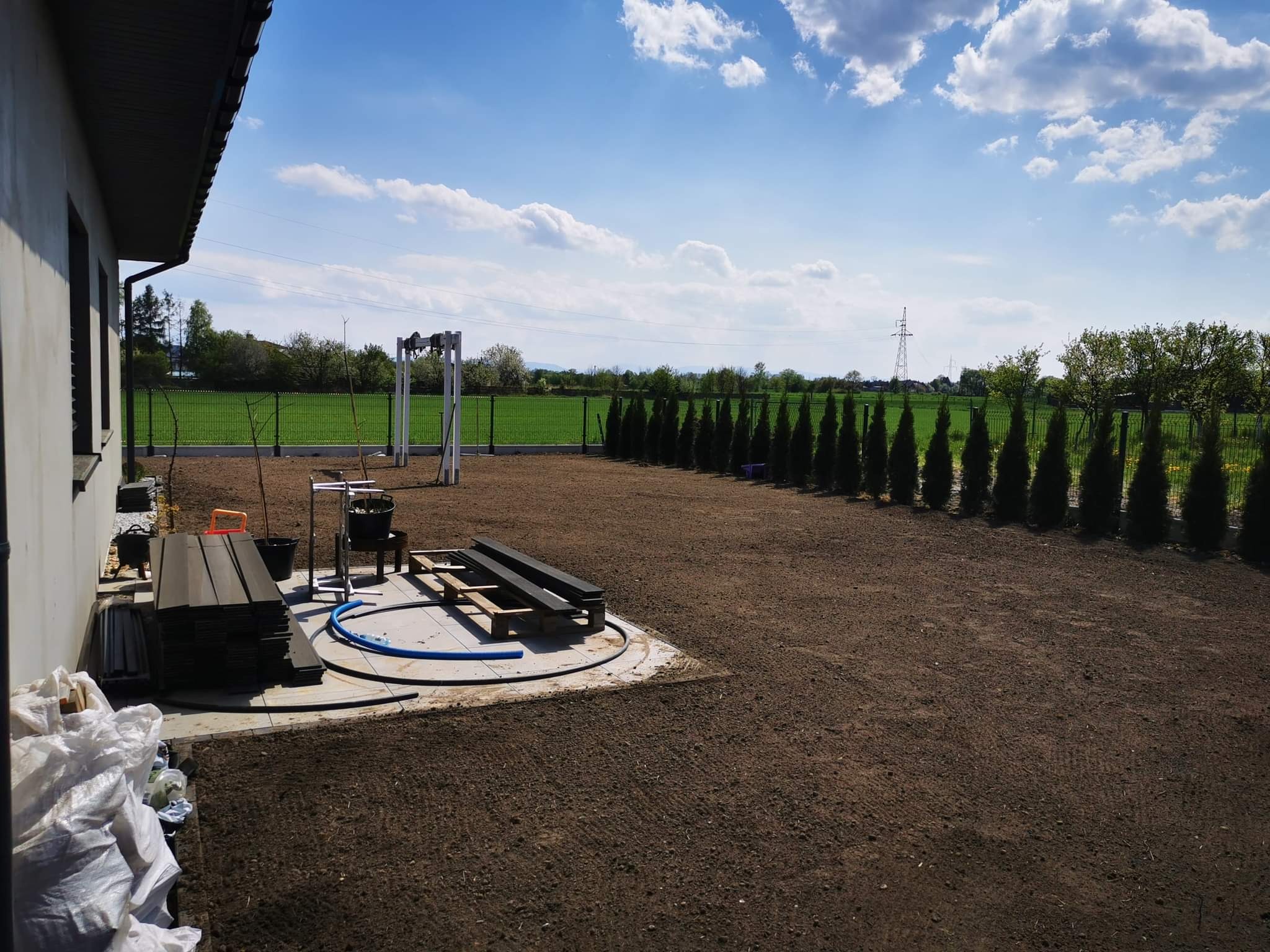Przygotowany teren pod trawnik z rolki, widoczne ułożone płyty tarasowe, ogrodzenie panelowe i rząd tuj, błękitne niebo z chmurami.