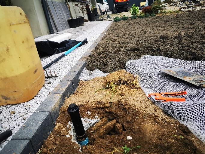 Instalacja zraszacza w ziemi przy obrzeżu trawnika, widoczna żółta beczka, grabie, łopata i nożyce ogrodowe oraz trawa z rolki w tle.