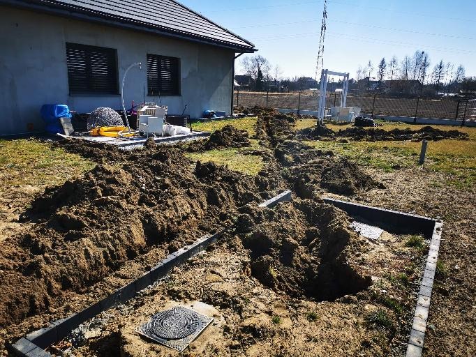Przygotowany wykop pod szambo z widocznymi obrzeżami z kostki brukowej, na tle domu z ciemnymi okiennicami i rozkopanym terenem.
