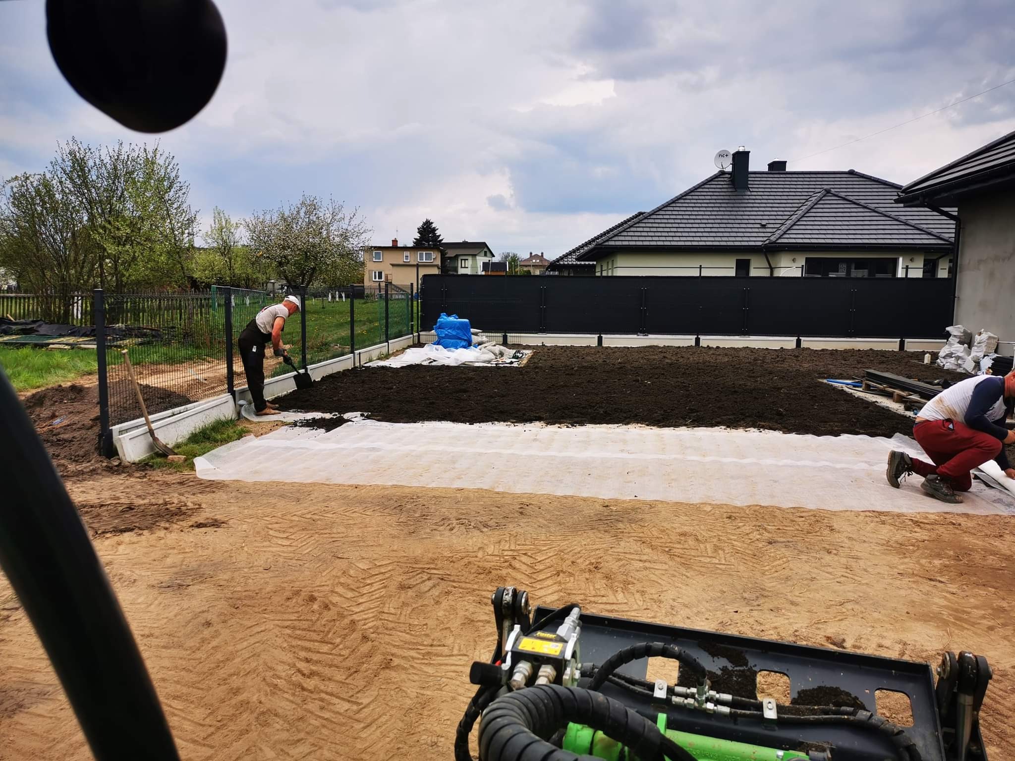 Przygotowanie terenu pod trawnik: pracownik wyrównuje ziemię łopatą, widoczna warstwa białej agrowłókniny, w tle ogrodzenie panelowe i zabudowa jednorodzinna.