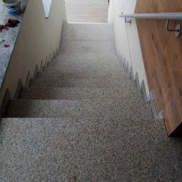 IDMbud - Granitowe schody zewnętrzne z drewnianą okładziną ściany po prawej stronie, widok z góry na dół, z widocznym wiadrem i narzędziami na górnym podeście.