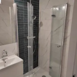 Nowoczesna kabina prysznicowa z czarnymi płytkami w strefie natrysku i białymi marmurkowymi płytkami na pozostałych ścianach, z umywalką w rogu, po remoncie.