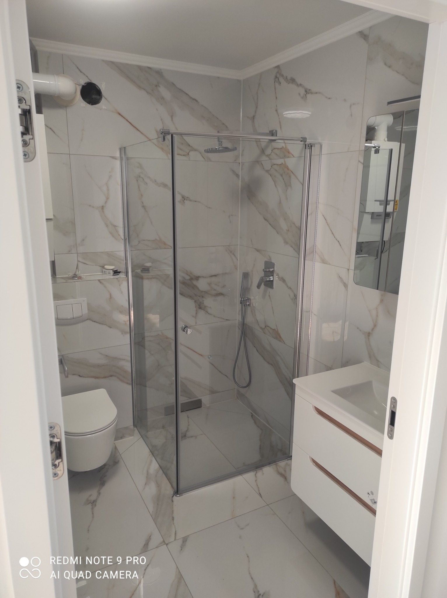 Wykończona łazienka z kabiną prysznicową, umywalką z szafką i toaletą, wykończona białymi płytkami imitującymi marmur, widok zza uchylonych drzwi.