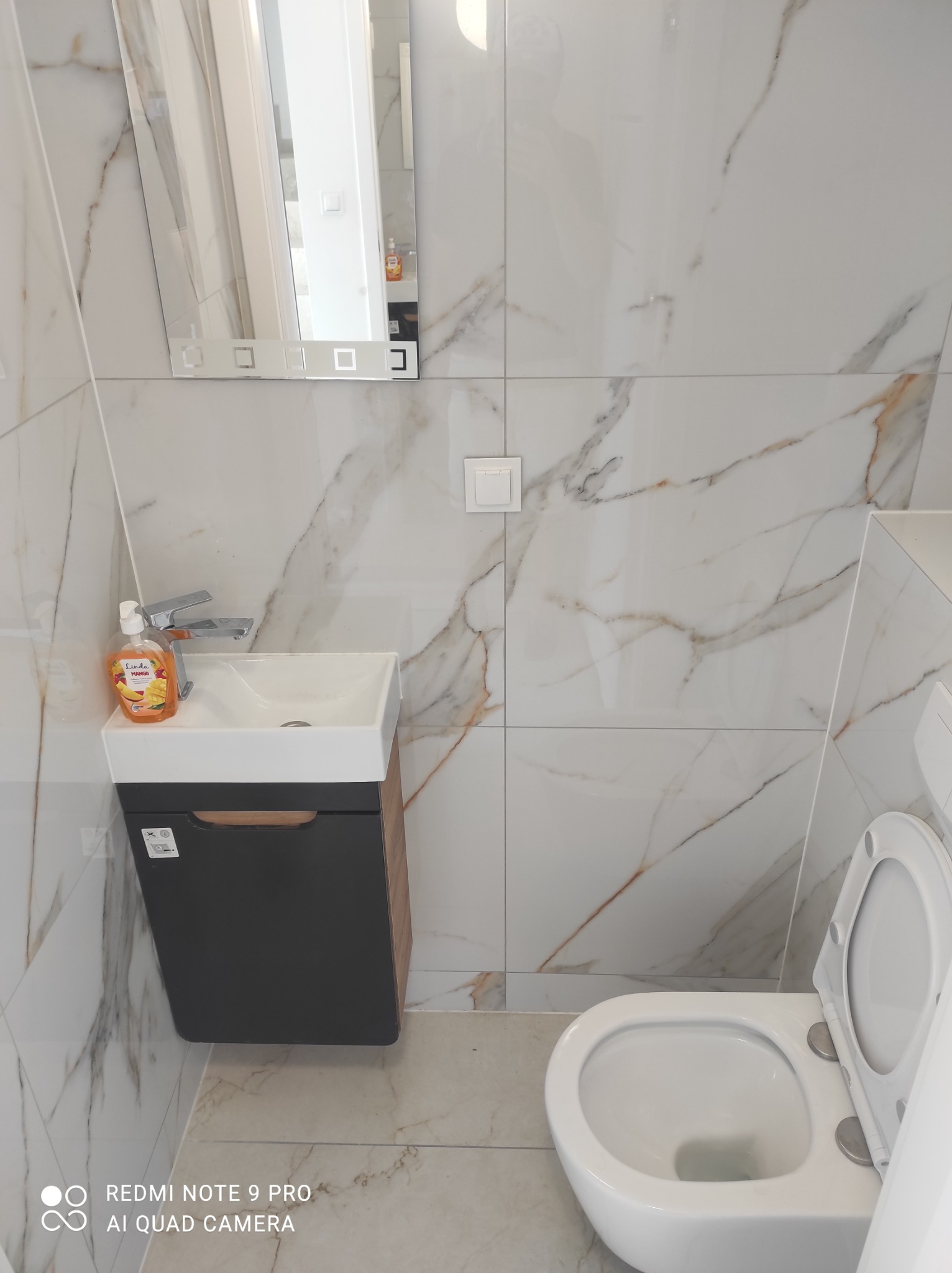 Biała łazienka z marmurowymi płytkami, umywalką z szafką w kolorze grafitowym i toaletą z otwartą klapą.