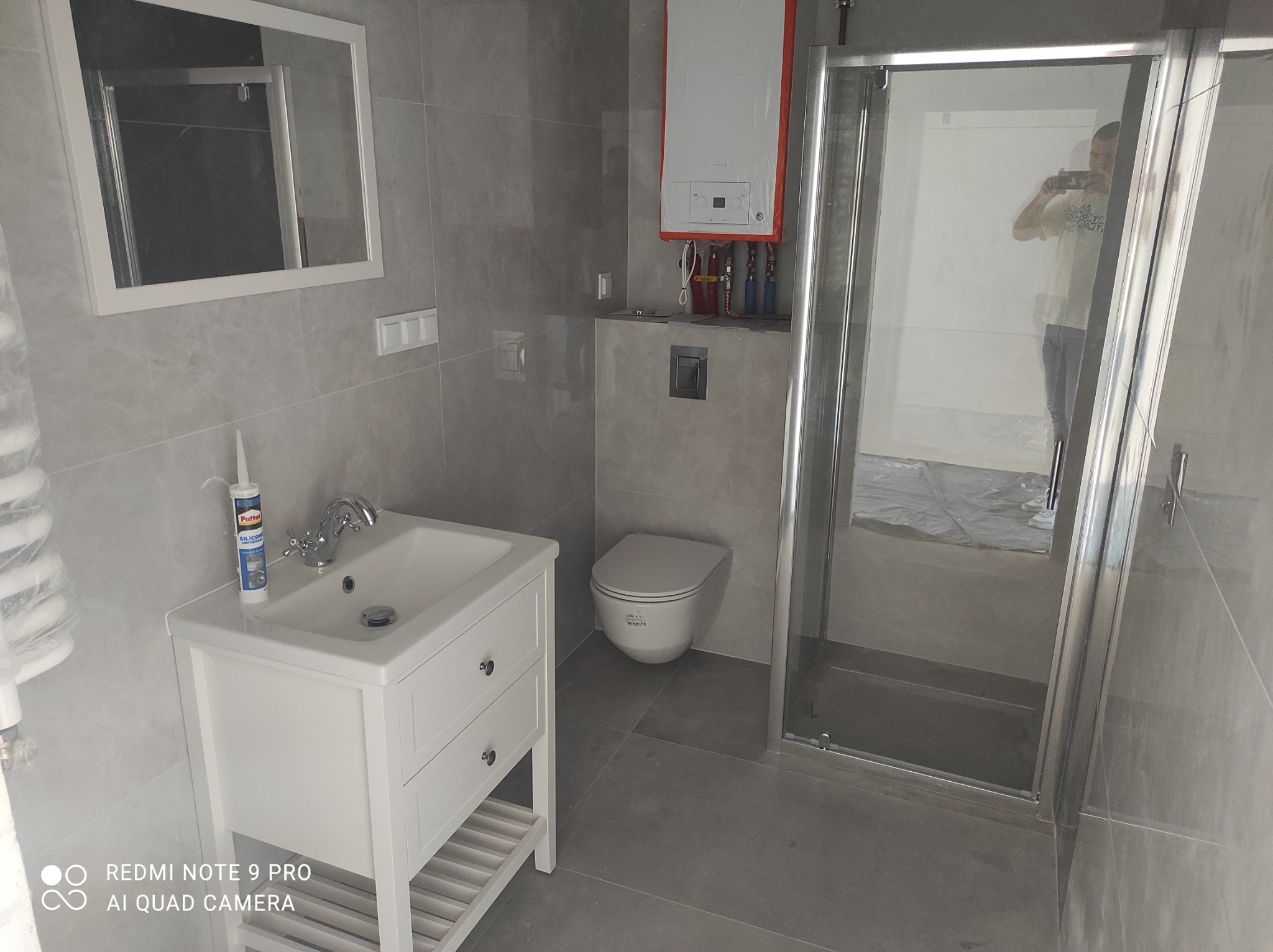 Szara łazienka po remoncie z białą umywalką na szafce, wiszącą toaletą i kabiną prysznicową; w lustrze odbija się osoba robiąca zdjęcie.