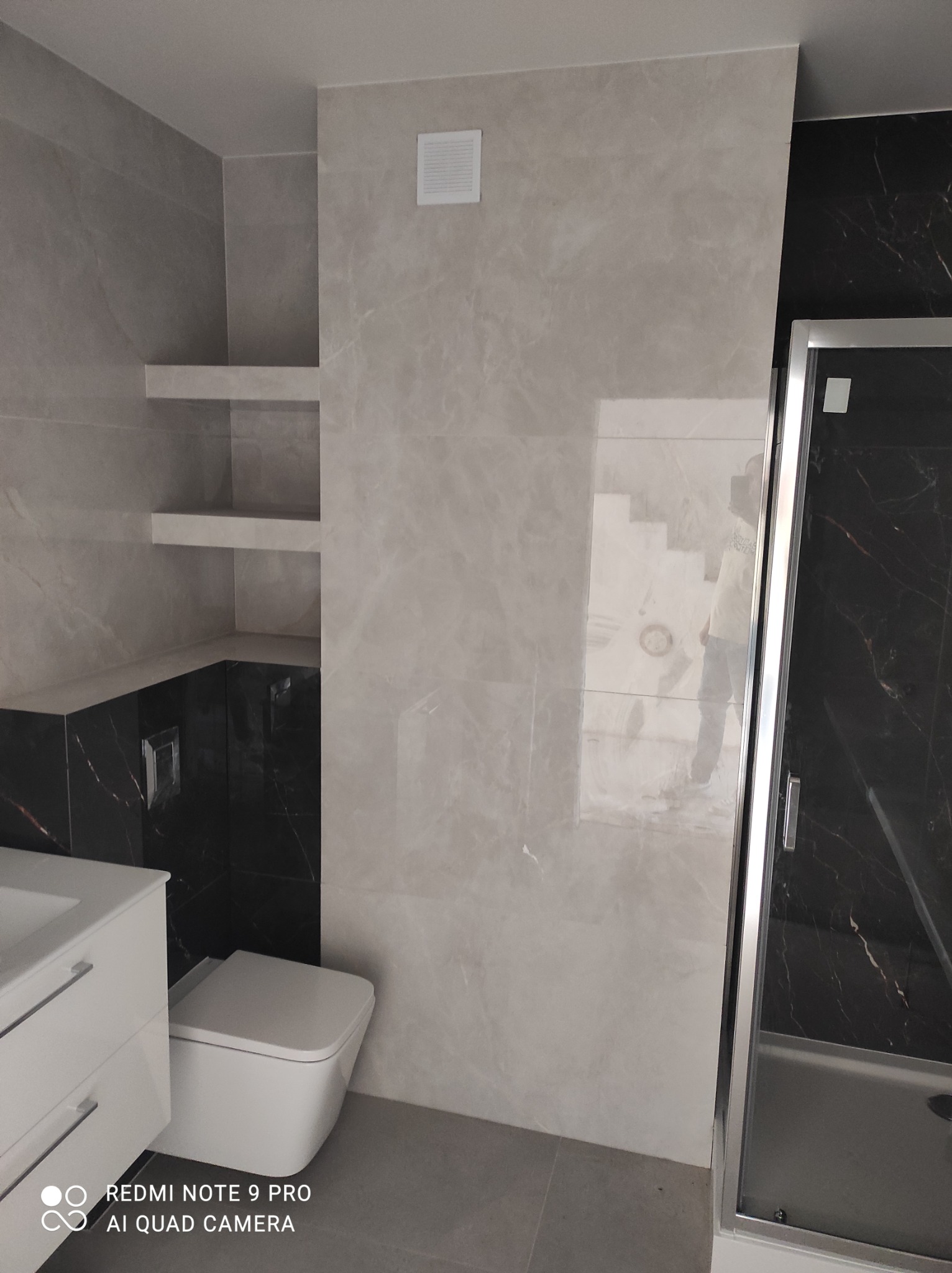 Wykończona łazienka z szarymi płytkami imitującymi marmur, podwieszana toaleta i szafka z umywalką, czarne płytki z białymi żyłami, oraz wnęka z półkami.