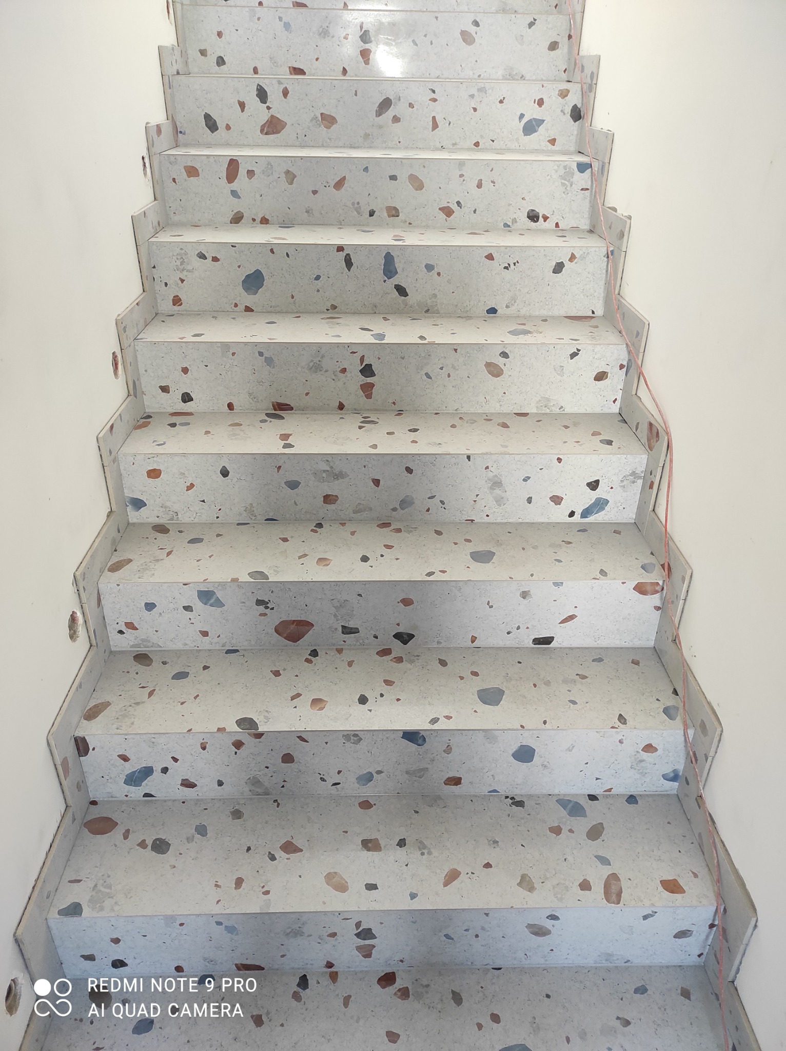 Schody wykończone płytkami lastryko w odcieniach szarości, beżu, brązu i niebieskiego, widok z dołu, z widocznymi śladami po wierceniu w ścianie.
