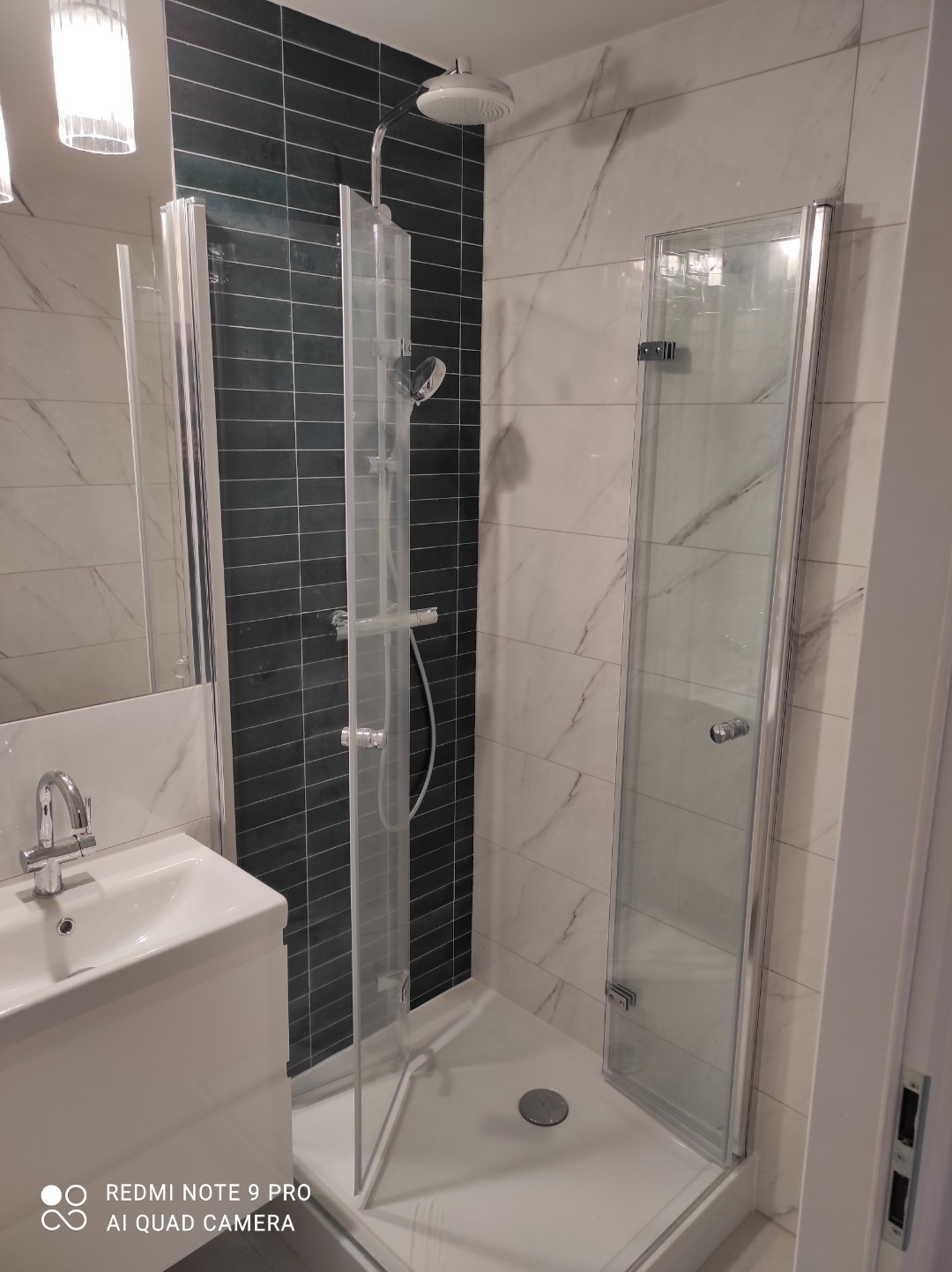 Nowoczesna kabina prysznicowa z czarnymi płytkami w strefie natrysku i białymi marmurkowymi płytkami na pozostałych ścianach, z umywalką w rogu, po remoncie.