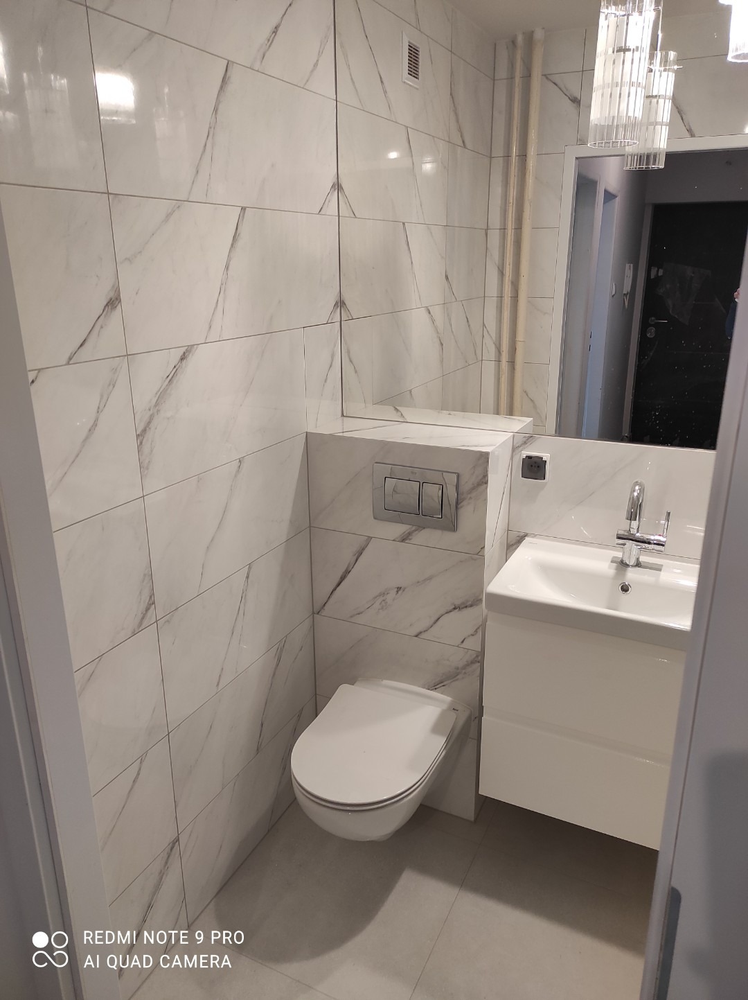 Nowoczesna, wykończona łazienka z białymi płytkami imitującymi marmur, podwieszana toaleta i umywalka z szafką, widoczne elementy armatury i oświetlenia.