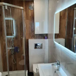 Nowoczesna łazienka z kabiną prysznicową, umywalką z szafką i lustrem z podświetleniem LED, wykończona płytkami imitującymi drewno i jasnymi płytkami ceramicznymi.