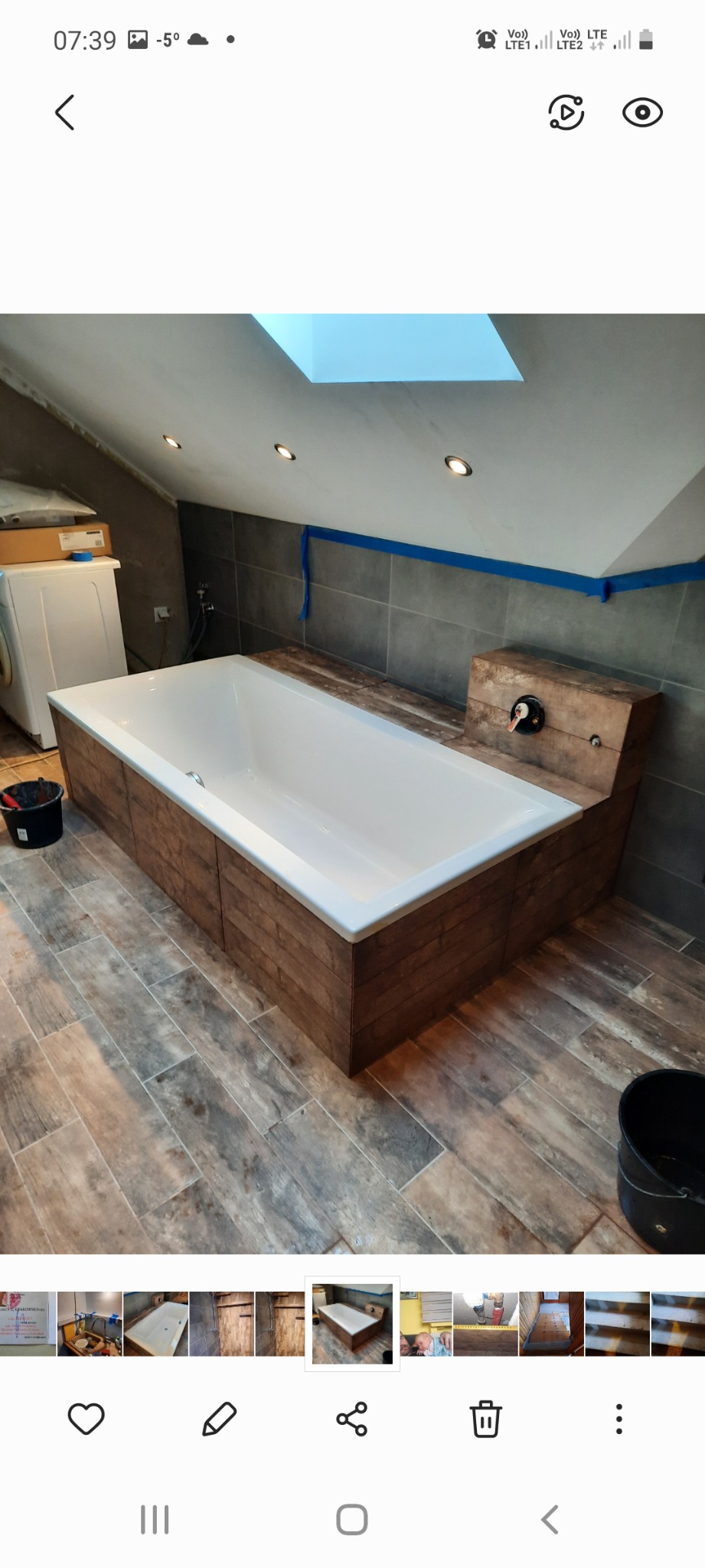 Biała wanna obudowana drewnopodobnymi panelami w łazience na poddaszu ze skośnym sufitem i oknem dachowym, widoczne płytki podłogowe imitujące drewno i szare płytki ścienne.