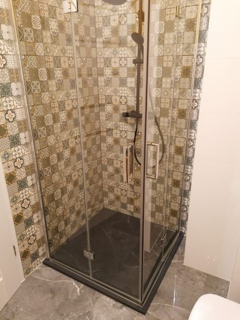 Szklana kabina prysznicowa z czarnym brodzikiem i mozaiką na ścianach, widok z boku, szare marmurowe płytki na podłodze, fragment toalety w rogu.