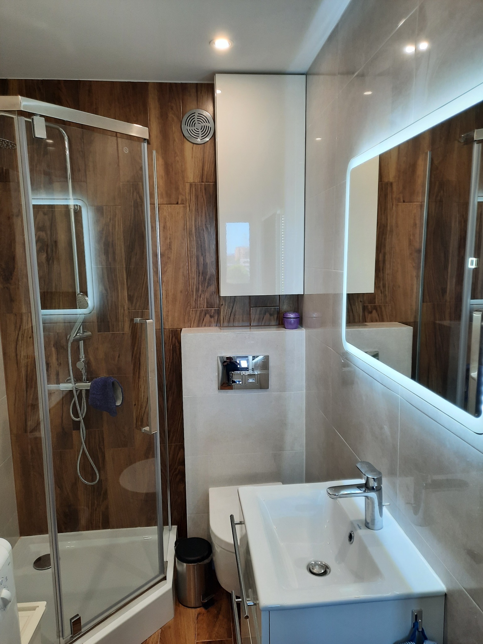 Nowoczesna łazienka z kabiną prysznicową, umywalką z szafką i lustrem z podświetleniem LED, wykończona płytkami imitującymi drewno i jasnymi płytkami ceramicznymi.