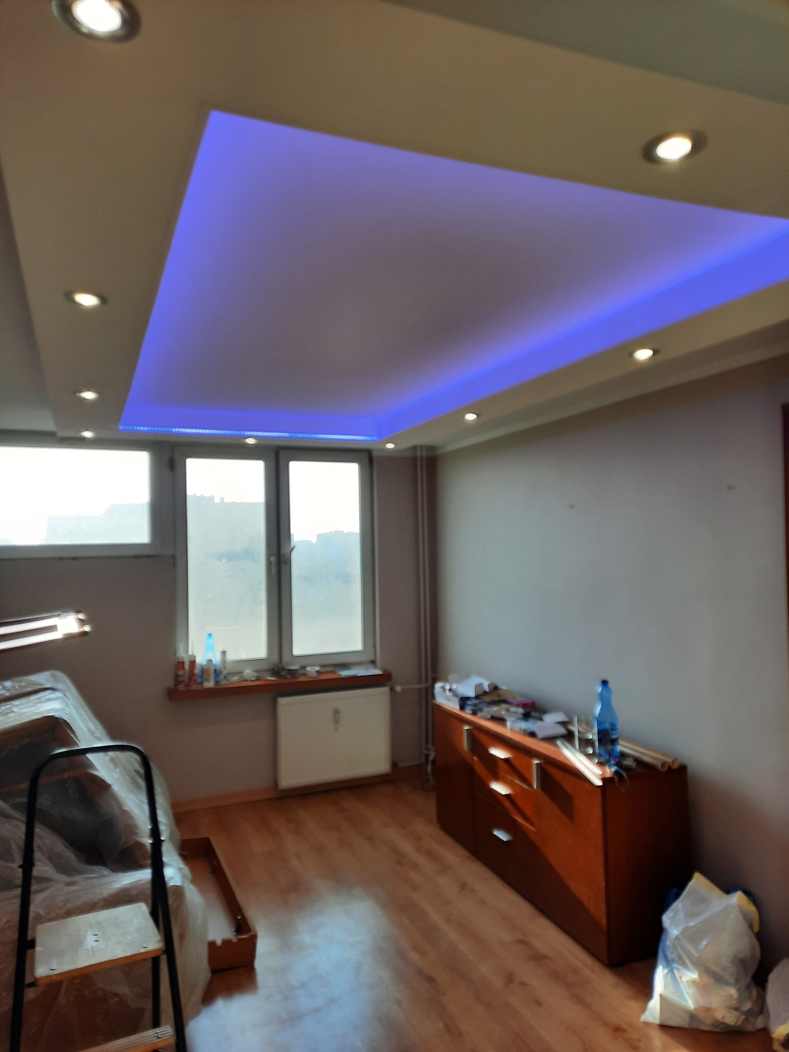 Sufit podwieszany z niebieskim oświetleniem LED w trakcie montażu, widoczna drabina i zabezpieczone meble folią malarską, na komodzie narzędzia i materiały budowlane.