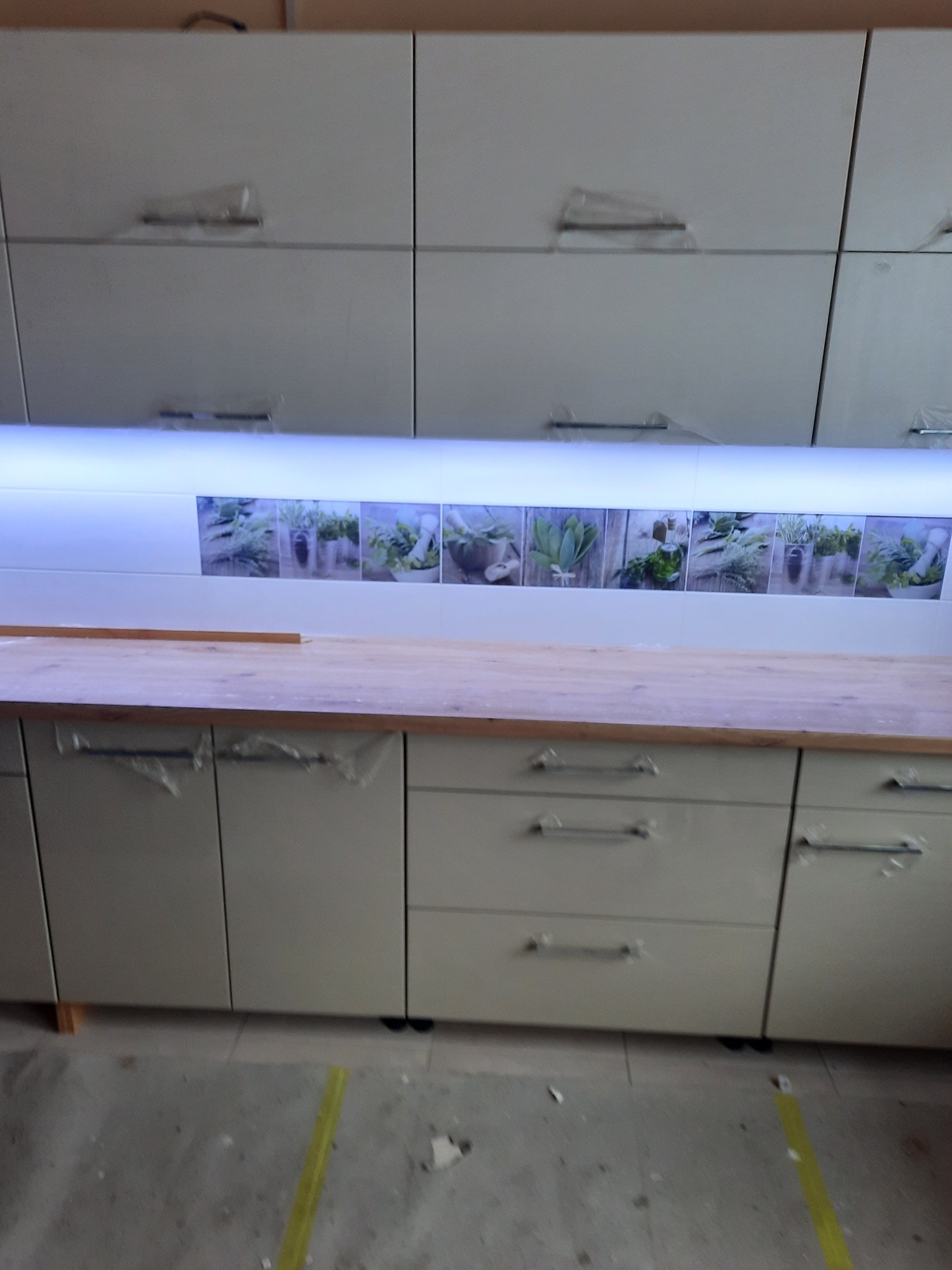 Niedokończona zabudowa kuchenna z białymi szafkami, blatem z jasnego drewna i panelem dekoracyjnym z motywem ziół nad blatem, osłonięte folią ochronną.