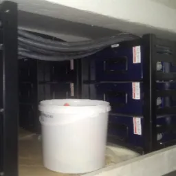 Rząd akumulatorów w szafie, widoczne kable i wiadro z farbą w pomieszczeniu.