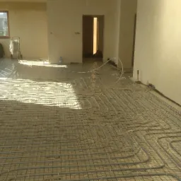 Rozłożona instalacja ogrzewania podłogowego na siatce w surowym wnętrzu, widoczne rury i folia ochronna.