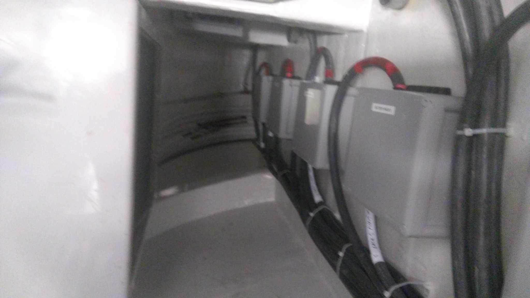 Szereg szarych skrzynek z kablami w białej obudowie, widoczne etykiety na kablach.